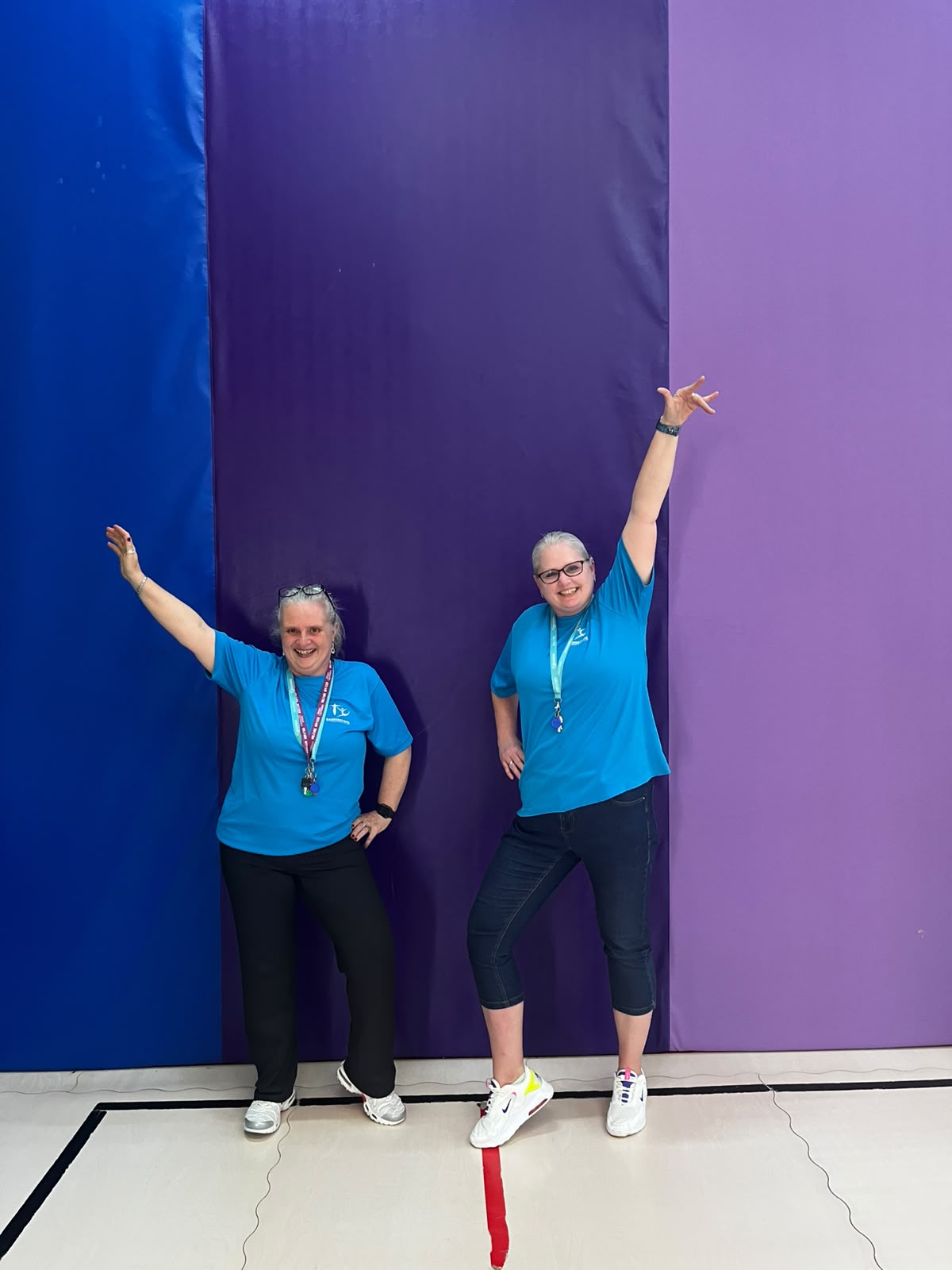 Two BGC Welfare Officers in club shirts with their arms raised, in front of the gym's crash mats